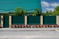 Green metal corrugated fence with brick pillars on the background of a beautiful house. Royalty Free Stock Photo