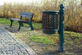 Green metal dustbin trash can stands on the grass nearby a metal bench with wood planks in the city park. Royalty Free Stock Photo