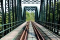 Green metal railway bridge construction with railway tracks surrounded with wooden sidewalk going towards overgrown forest vegetat