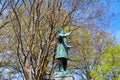 A green metal statue of a man in a overcoat surrounded by bare winter trees and lush green trees with blue sky