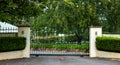 Green metal wrought iron driveway property entrance gates set in concrete fence, garden shrubs, trees, lights