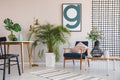 Green plant in white pot between wooden table with leaf in black vase and retro armchair with pastel pink pillow, real photo with