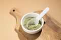 Green powder in a small white ceramic bowl, with a white spoon on a wooden board, on a beige background.