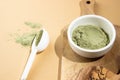 Green powder in a small white ceramic bowl, with a white spoon on a wooden board, on a beige background