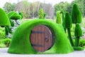 Green sculpture of small house with round wooden opened door surrounded by mushrooms made from artificial grass. Urban