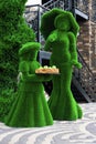 Green sculptures of young girl and a lady with umprella in long dress made from artificial grass. Girl holds wooden tray