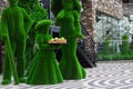 Green sculptures of young lady, man and woman with umprella made from artificial grass. Girl holds wooden tray with