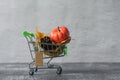 Green small toy shopping cart with birch leaves, cone and pumpkin on a concrete wall background. Price label note with rope for Royalty Free Stock Photo