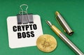 On a green surface, a bitcoin coin, a pen and a sheet of paper with the inscription - Crypto boss