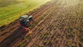 Green tractor with plougher driving through bean plantation next to lush green field and faded tree silhouettes in the