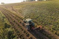 Green tractor with plougher driving through bean plantation next to lush green field and faded tree silhouettes in the