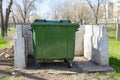 A green trash can stands on a site with a white concrete fence in a public park. Garbage collection and disposal using a plastic