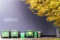 View of green garbage containers placed against a gray wall and yellow tree foliage next to them Royalty Free Stock Photo