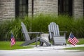 Two grey Adirondack chairs sitting on a small patio with table between them in front of tall decorative grass and windows of rock