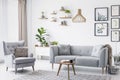 Grey armchair next to settee in bright living room interior with posters and wooden table. Real photo Royalty Free Stock Photo