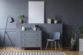 Grey cabinet between black lamp and chair in living room interior with mockup of poster. Real photo