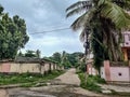 Grey color road constructed from cement surrounded with coconut trees and Indian styled homes in cloudy climate. No people around