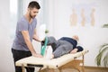 Grey elderly man lies on a table, and young doctor helps him during exercises