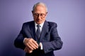 Grey haired senior business man wearing glasses and elegant suit and tie over purple background Checking the time on wrist watch, Royalty Free Stock Photo