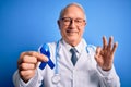 Grey haired senior doctor man holding colon cancer awareness blue ribbon over blue background doing ok sign with fingers, Royalty Free Stock Photo