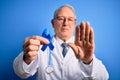 Grey haired senior doctor man holding colon cancer awareness blue ribbon over blue background with open hand doing stop sign with Royalty Free Stock Photo