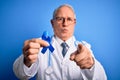 Grey haired senior doctor man holding colon cancer awareness blue ribbon over blue background pointing with finger to the camera Royalty Free Stock Photo