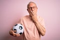 Grey haired senior football player man holding soccer ball over pink isolated background cover mouth with hand shocked with shame