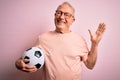 Grey haired senior football player man holding soccer ball over pink isolated background very happy and excited, winner expression