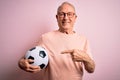 Grey haired senior football player man holding soccer ball over pink isolated background very happy pointing with hand and finger