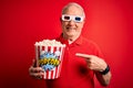 Grey haired senior man wearing 3d movie glasses and eating popcorn over red background very happy pointing with hand and finger Royalty Free Stock Photo