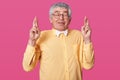 Grey haired senior raises hands up and crosses fingers. Elderly man with fashion yellow shirt poses in studio isolated over rose Royalty Free Stock Photo