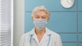Grey haired senior woman doctor wearing surgical mask and white robe with stethoscope looks into camera