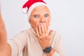 Grey-haired woman wearing Crhistmas Santa hat make selfie over isolated white background cover mouth with hand shocked with shame Royalty Free Stock Photo