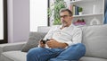 Grey-haired young hispanic man in glasses, beard, and serious expression immersed in intense online gaming, using joystick, Royalty Free Stock Photo