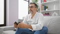 Grey-haired young hispanic man in glasses, beard, and serious expression immersed in intense online gaming, using joystick, Royalty Free Stock Photo