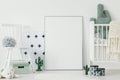 Grey pompom placed on white chair standing in bright baby bedroom interior with cactus shaped decor and empty poster on the floor