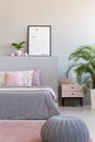 Grey pouf next to bed with cushions in modern bedroom interior with poster and plants. Real photo Royalty Free Stock Photo