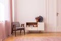 Grey vintage armchair next to wooden cabinet with books and heather in pot, real photo with copy space on the empty wall