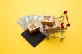 Grey wallet with hundred dollar bills, three delivery boxes and shopping trolley on yellow background