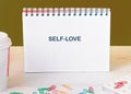 A grid-pattern notebook displays the phrase self love, surrounded by colorful paper clips and a coffee cup on a