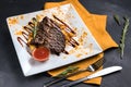 Grilled beef steak with potatoes, sauce and spices on white plate. Spoon, fork and orange tablecloth on dark gray texture