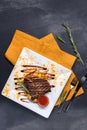 Grilled beef steak with potatoes, sauce and spices on white plate. Spoon, fork and orange tablecloth on dark gray texture