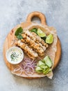 Grilled chicken on metal skewers on spinach leaves, slices cucumber, red onion and lime and sauce in a bowl on wood board