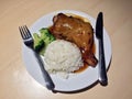 Grilled chicken roast topped with sauce and boiled broccoli and white cooked rice with Knife and fork in the white dish.