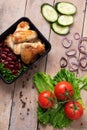 plastic container with grilled chicken wings and raw vegetables on rustic background