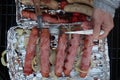 Grilled sausages in ham roll on thin perforated aluminium plate, turned with fork and kitchen knife. Some other sausages.