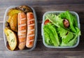 Grilled sausages, potatoes, green lettuce with tomatoes in a plastic box. Unhealthy diet. Wooden background Royalty Free Stock Photo