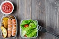 Grilled sausages, potatoes, green lettuce with tomatoes in a plastic box. Unhealthy diet. Wooden background. Copy space Royalty Free Stock Photo