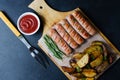Grilled sausages on a wooden chopping Board. Fried potatoes, rosemary, tomato ketchup. Unhealthy diet. Dark background Royalty Free Stock Photo