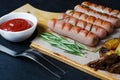 Grilled sausages on a wooden chopping Board. Fried potatoes, rosemary, tomato ketchup. Unhealthy diet. Dark background Royalty Free Stock Photo